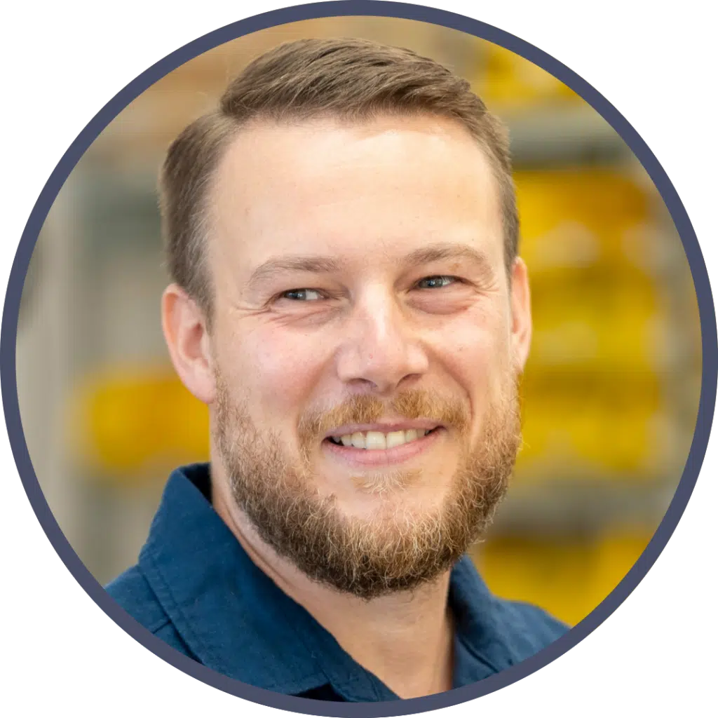 Team: Tobias Sebastian Hombach ist Gesellschafter in der Wurm & Hombach GbR Porträt eines Mannes mit Bart, der ein dunkelblaues Hemd trägt und in die Ferne lächelt. Im Hintergrund sind gelbe Strukturen zu erkennen.