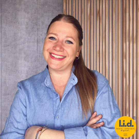Lea Wurm lächelt mit verschränkten Armen vor einer Wand aus Holzlamellen und Betonoptik, mit l.i.d.-Logo unten rechts.