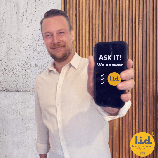 Tobias Hombach zeigt ein Smartphone mit „ASK IT. We answer.“ und dem l.i.d.-Logo in die Kamera.